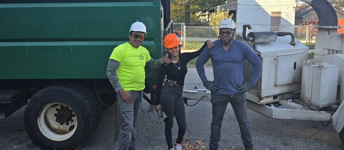 Three crew members standing in front of a green truck and a wood chipper on a leaf-covered street.